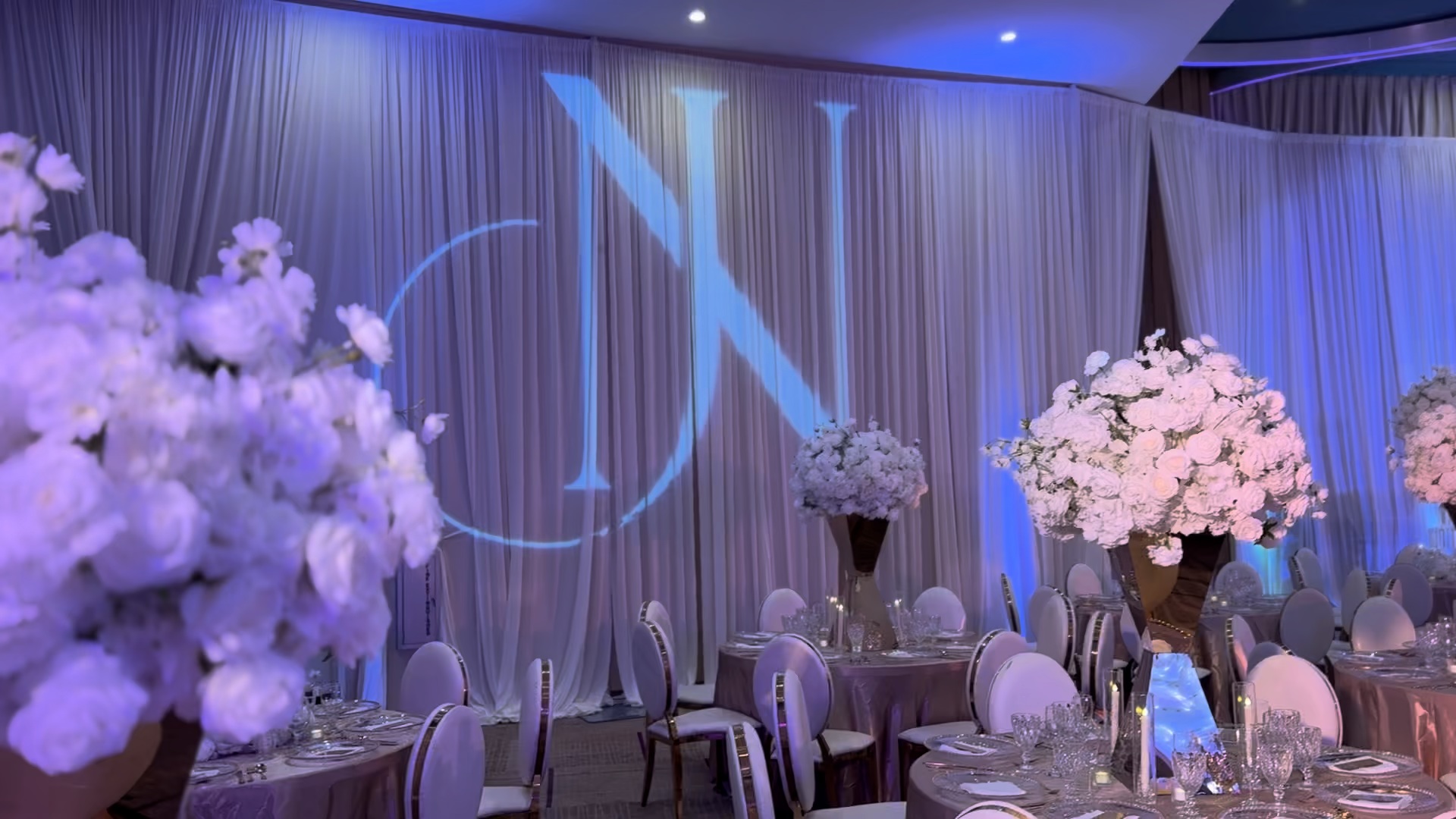 Large custom monogram gobo projected onto ballroom draping, framed by white floral centerpieces