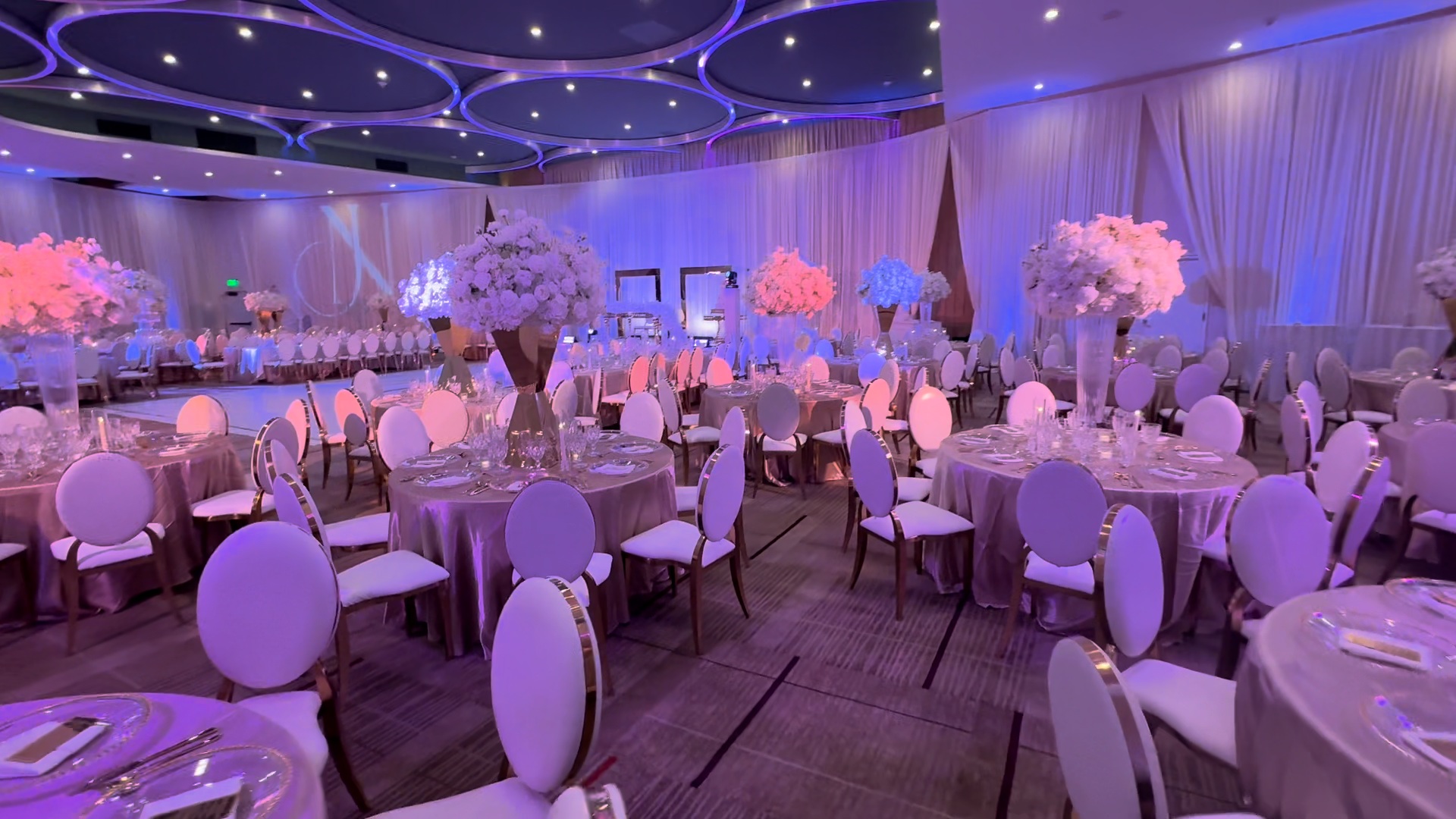 Ballroom reception with rows of floral centerpieces, lit draping, and formal table settings