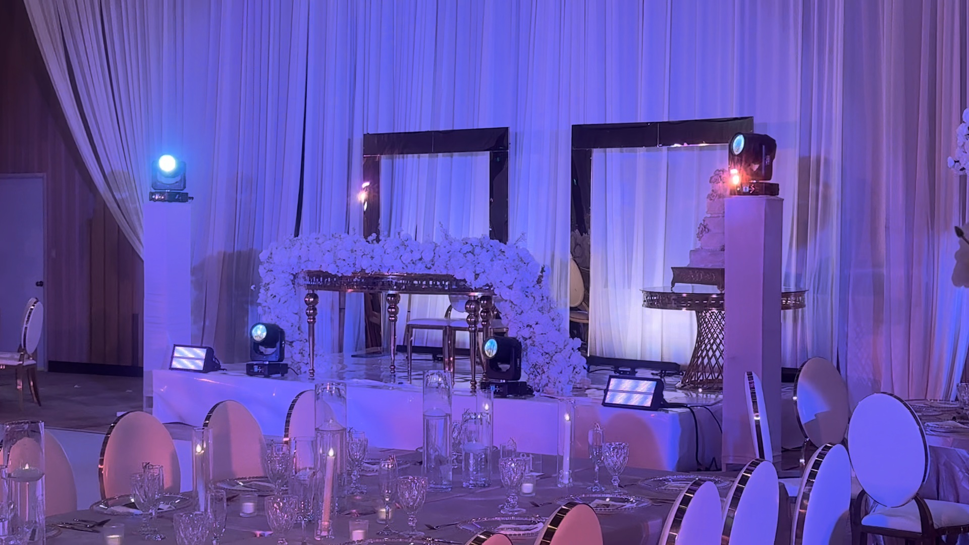 Sweetheart table on a raised white stage with flowers, two mirror-frame backdrops, moving heads, and a cake table